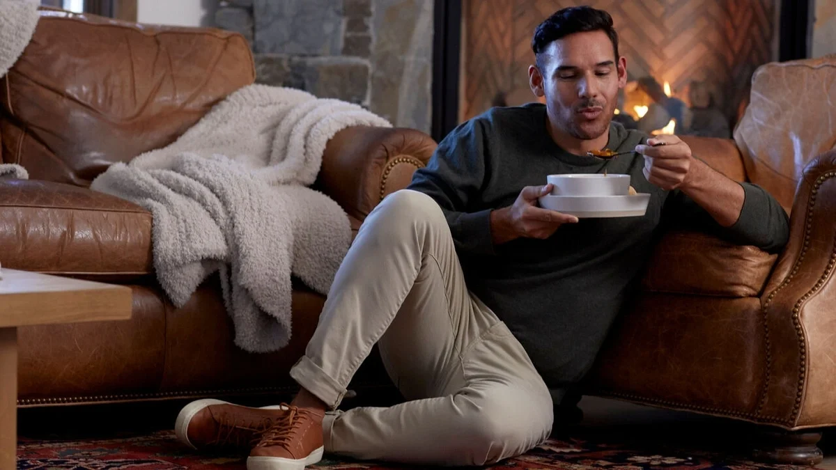 Person sitting on the floor beside a couch in front of a fireplace, enjoying a bowl of soup with a cozy throw blanket draped over the sofa nearby.