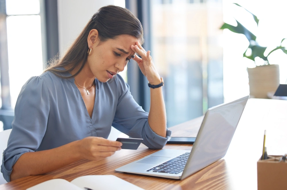 Woman sat at a laptop and looking at her credit card with a concerned expression