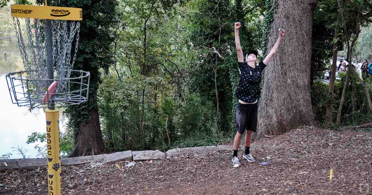 Young man holds up his hands in triumph near an elevated disc golf basket