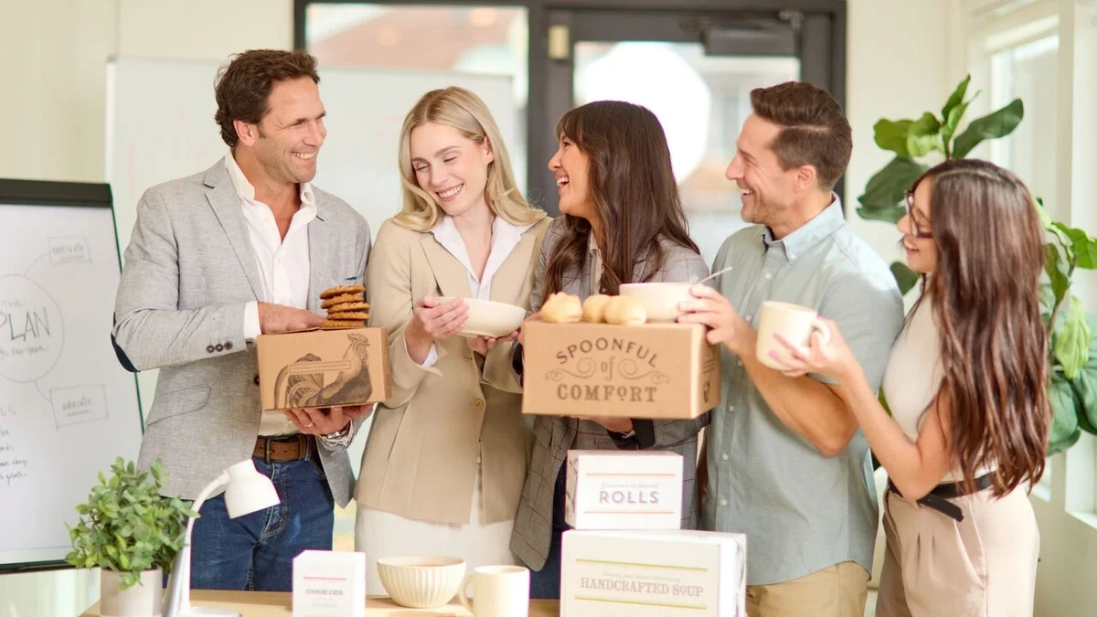 Five coworkers standing together in an office, smiling and sharing bowls of soup, cookies, and Spoonful of Comfort gift boxes during a group gathering.