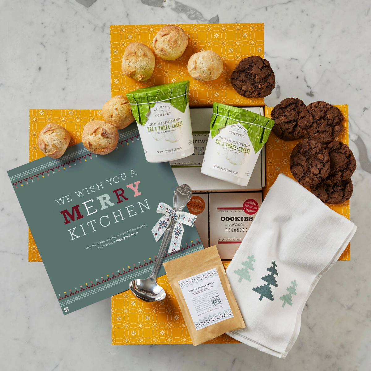A sign that says "We Wish You a Merry Kitchen" sits next two a couple packages of soup, 6 cookies, a ladel, a kitchen towel, a package of mulled simmer spices and two sets of 3 rolls.