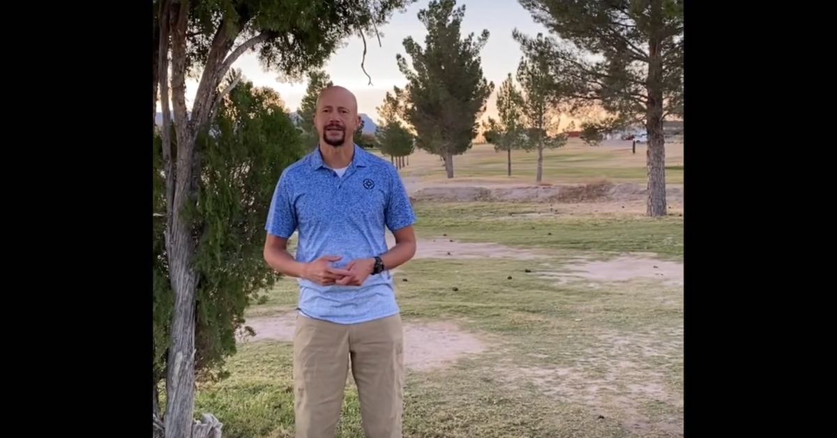 A man in khakis and a blue collared tee shirt stands on a slightly run down golf course