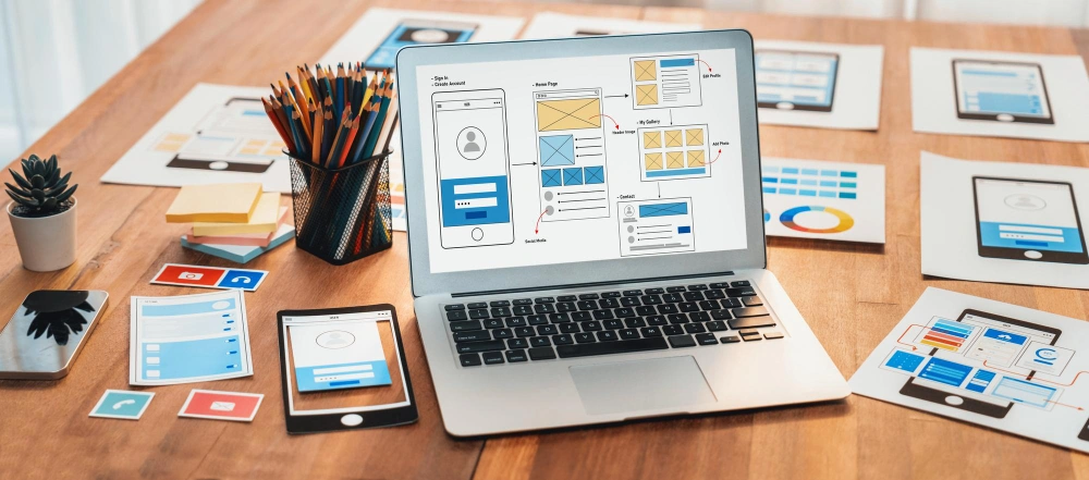 A photo of a laptop with user interface designs on the screen surrounded by various digital devices and notes, illustrating a user experience design process.