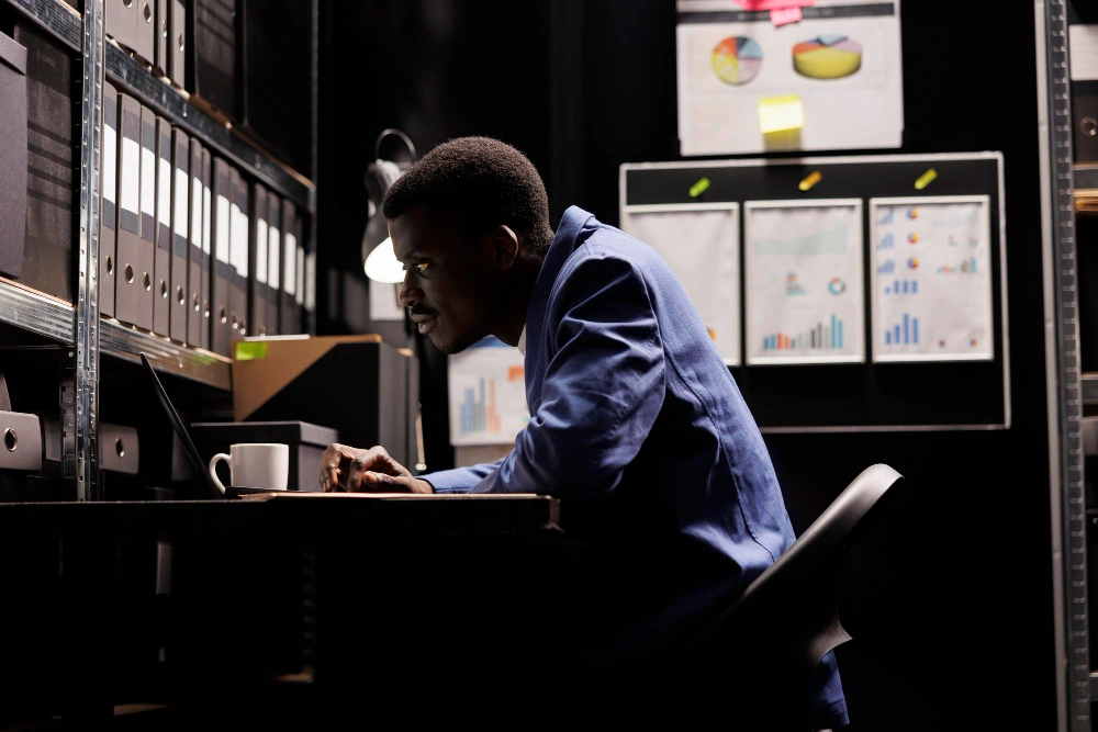 A man working late at night in a dimly lit office, surrounded by shelves filled with binders and charts on the wall.