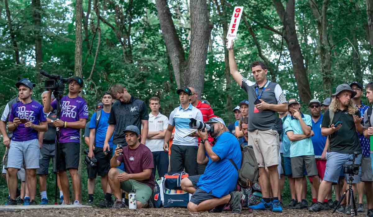 A group of people at a disc golf event wait in a group in the woods