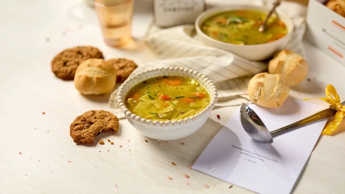 Close-up of a bowl of chicken soup with rolls and cookies, alongside a metal spoon resting on a handwritten note, with another bowl of soup in the background.