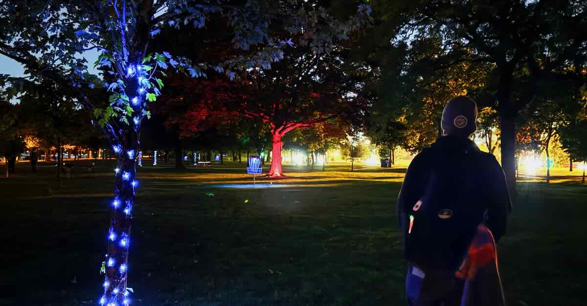 A man stands with disc golf equipment near a tree lit up with blue lights
