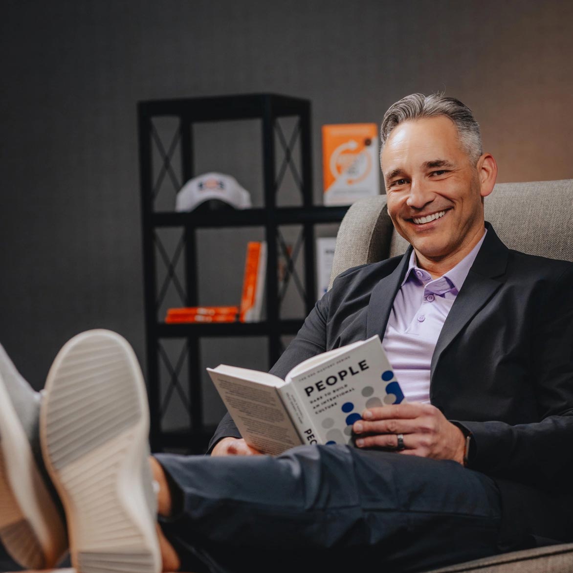 Portrait of Mark O'Donnell sitting on a chair with his feet up reading a book