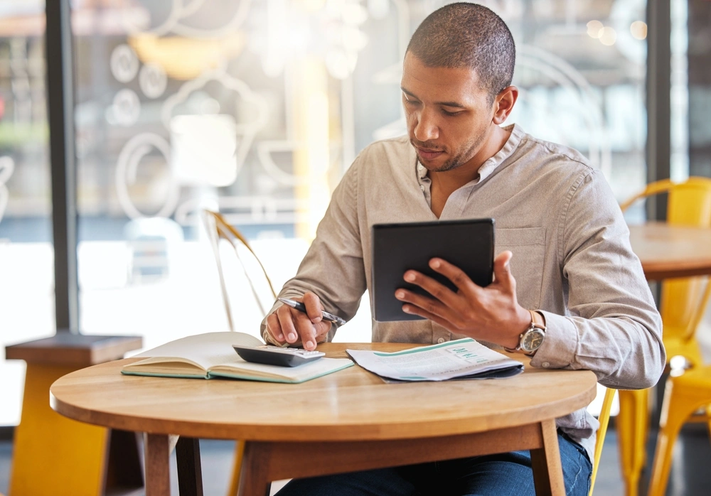 man sat at a table in a cafe looking at his laptop and calculator