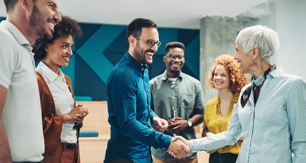 A group of professionals in a modern office shares a friendly moment, with two people in the foreground shaking hands and others smiling around them.