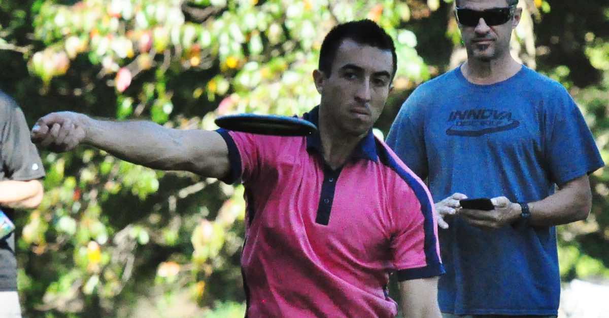 Man in pink shirt throwing a disc golf disc. A man in a blue shirt looking on is visible behind