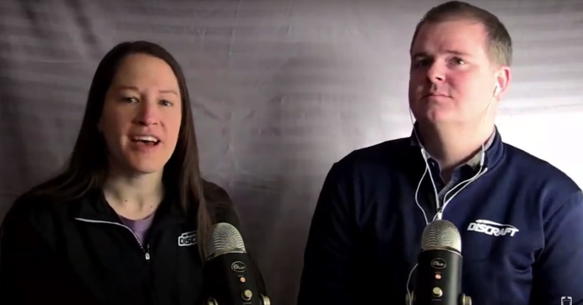 A woman and a man speak into microphones in a dim room in front of a bed sheet
