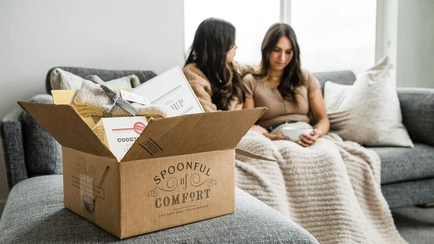 Open Spoonful of Comfort gift box filled with comfort items in the foreground, while two women sit together on a couch under blankets in the background, sharing a quiet moment.