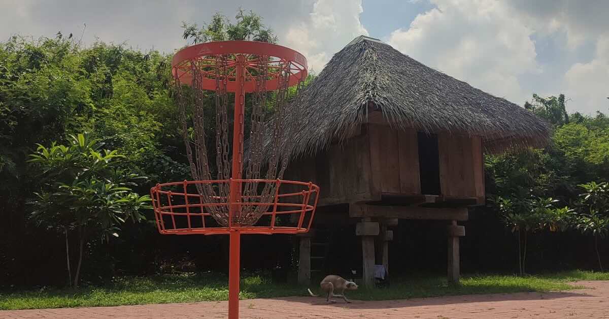 Orange disc golf basket in front of a building with a palm frond roof