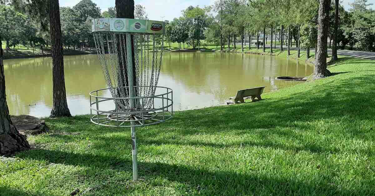 A disc golf basket in front of a pond wth a bench and mown grass