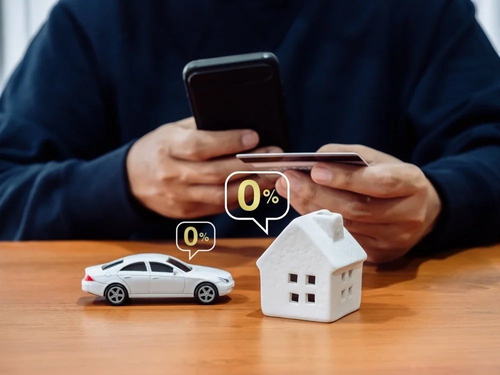 Close up of a man with his phone and credit card with a model home and a model car in front of him with 0% bubbles around the models