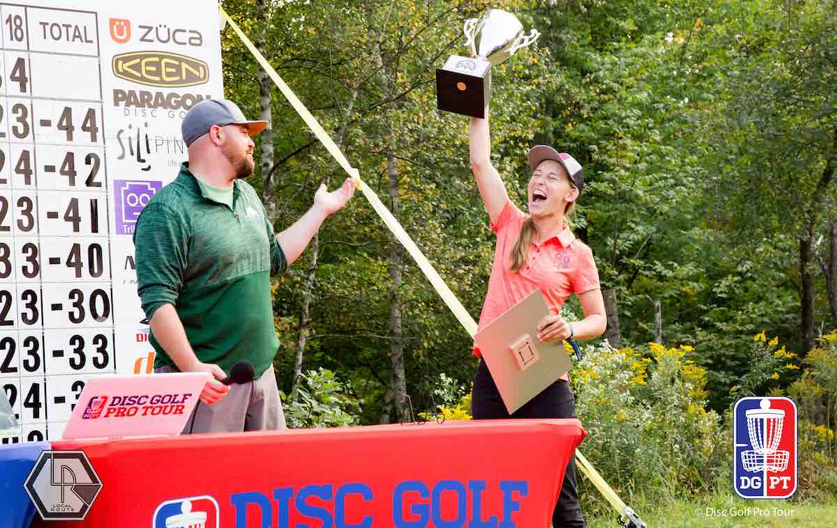 A man with a beard and a young woman stand at a table with a scoreboard behind them. The young woman is holding a trophy and shouting in joy