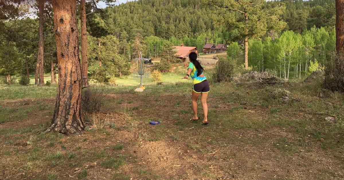 A woman in tye-dye shirt and black running shorts putting at a disc golf basket in wooded, mountainous area