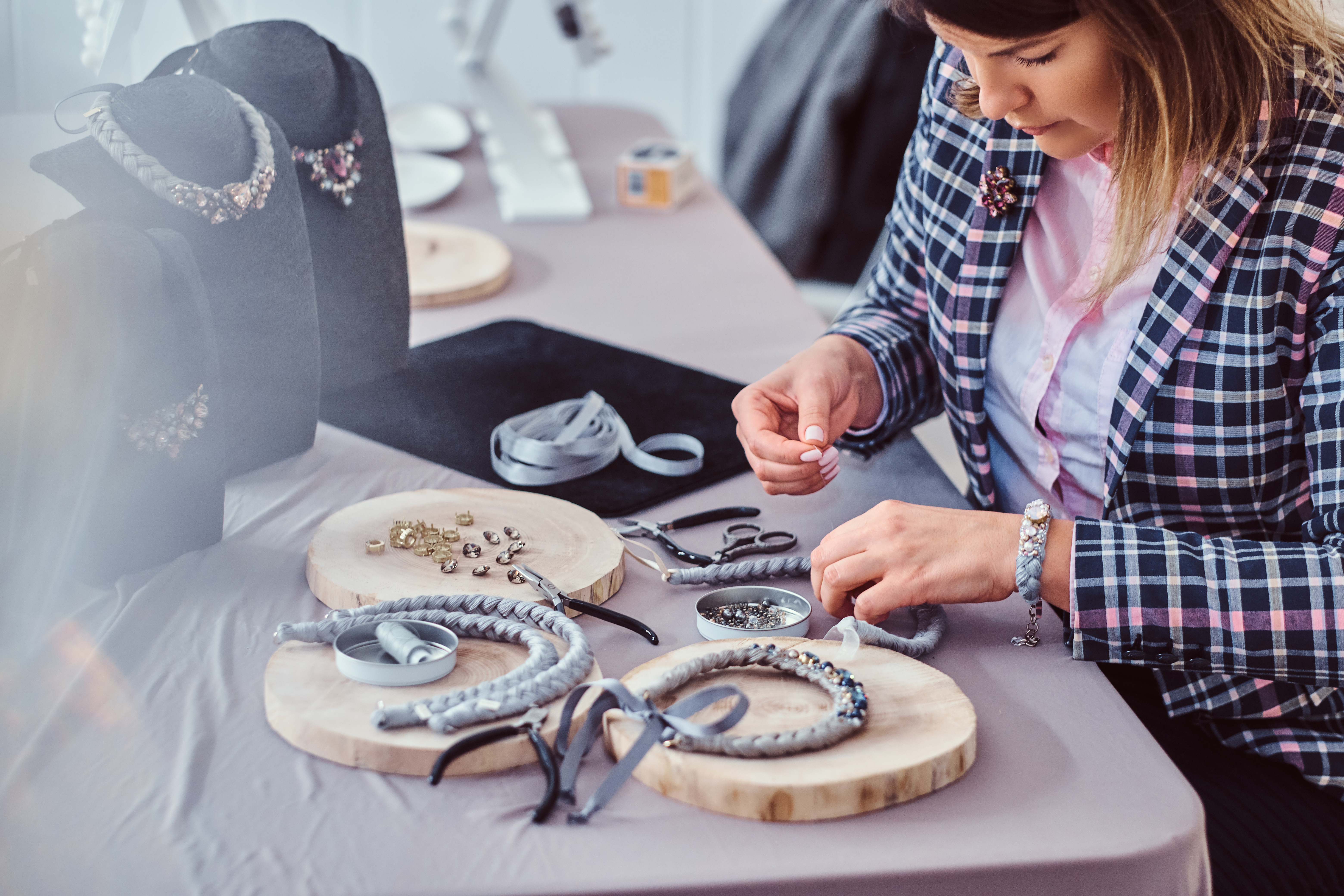 woman in a plaid jacket making jewelry with ribbon and beads surrounded by finished jewelry