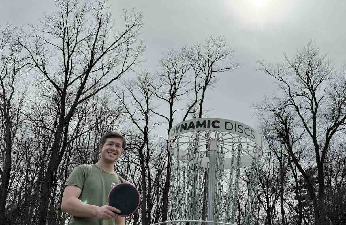 A man poses with a disc in front of a disc golf basket