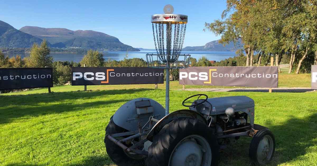A disc golf basket is mounted on a tractor on a well-mown green and there's a fjord in the background