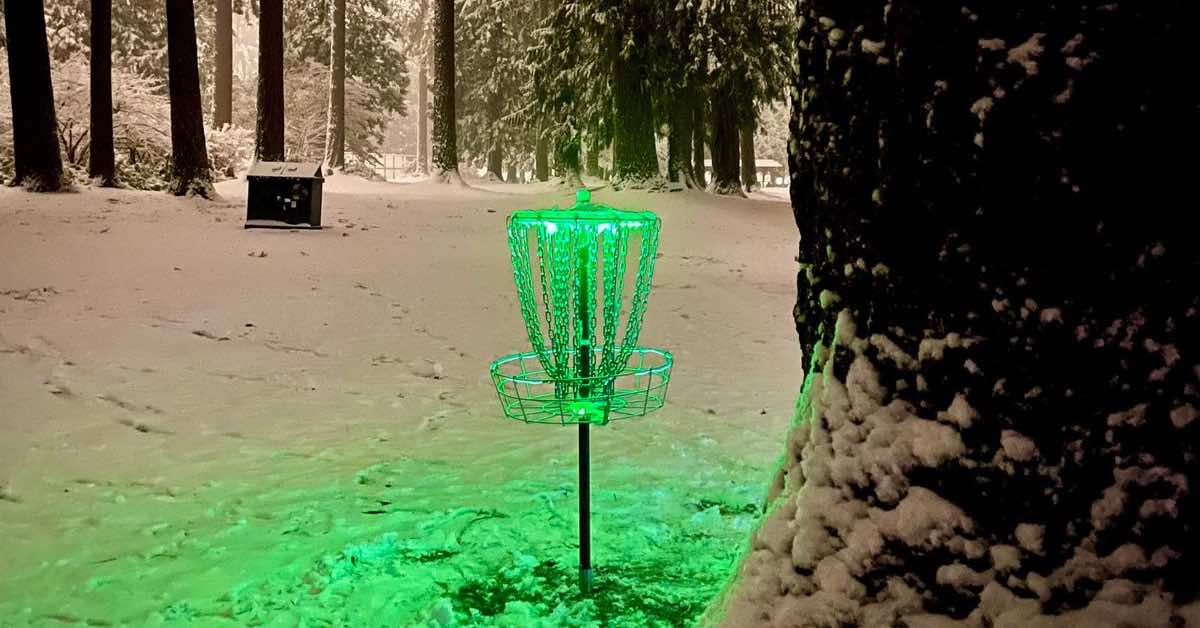 A disc golf basket in snowy woods at night lit by a green light