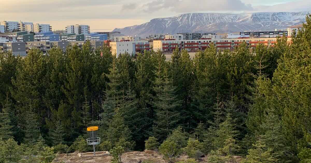 Disc golf basket in a mountainous landscape with snow-capped peaks and apartments in the distance