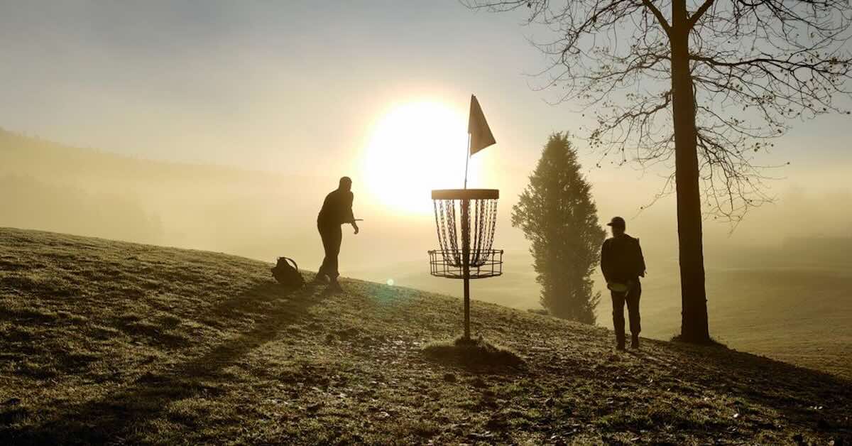 Two people playing disc golf to a basket on a hill with morning light streaming through mist behind them