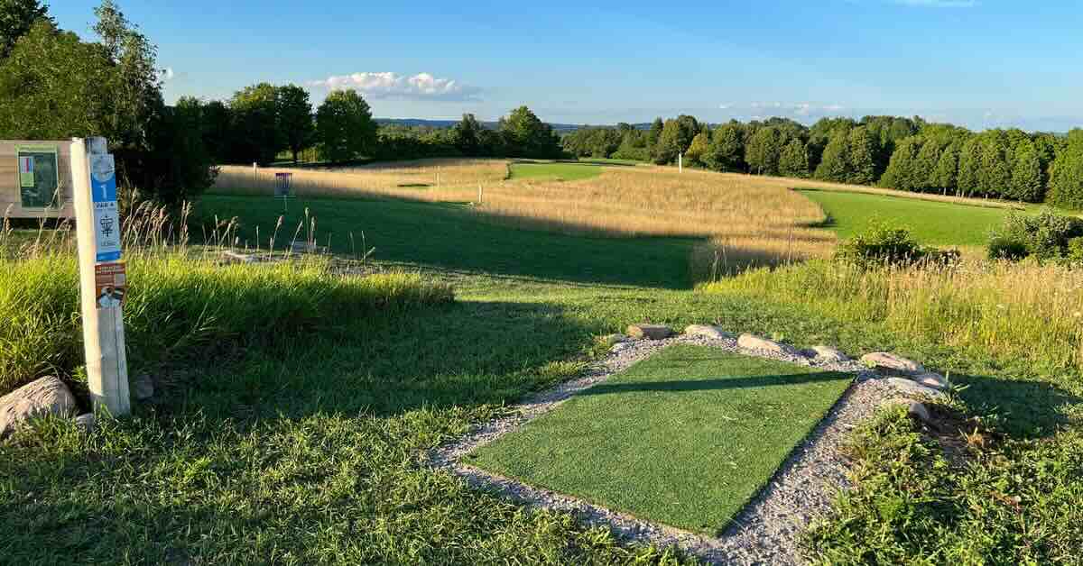 A turf disc golf tee pad in a well-mown open area with hills and trees in distance