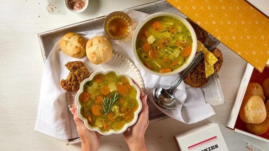 Overhead view of a tray with two bowls of chicken soup, fresh rolls, cookies, a glass of tea, and a metal ladle with a yellow ribbon, with one bowl being held in both hands.