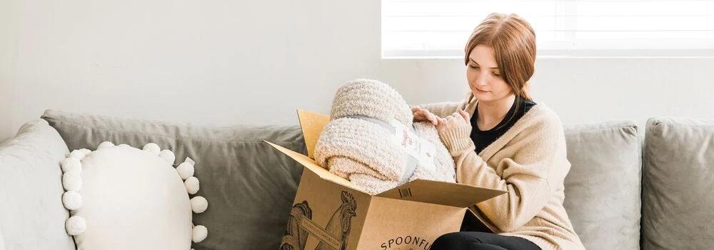 A female sitting on a couch unboxing a blanket from a package.