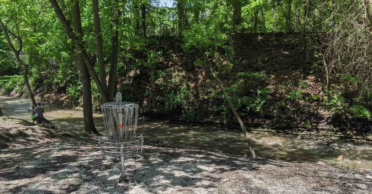 A disc golf basket on a pebbly bank near a flowing stream
