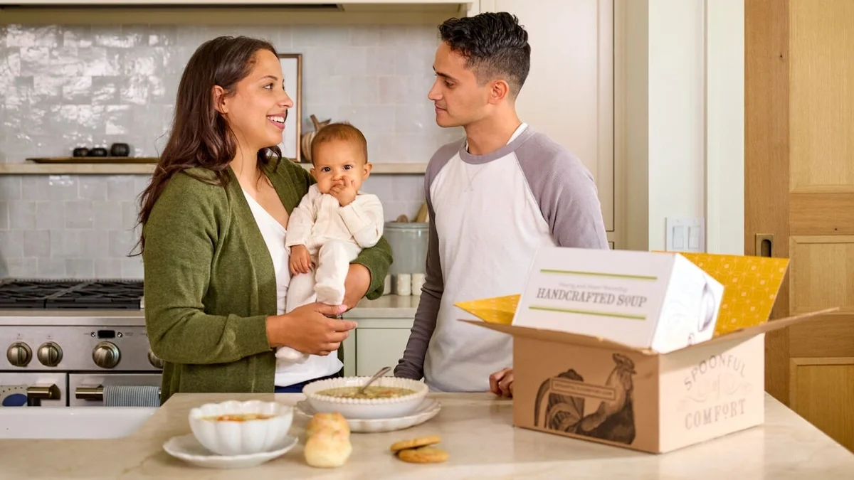 Couple standing in a kitchen with their baby, smiling beside an open Spoonful of Comfort gift box, with bowls of soup, rolls, and cookies on the counter.