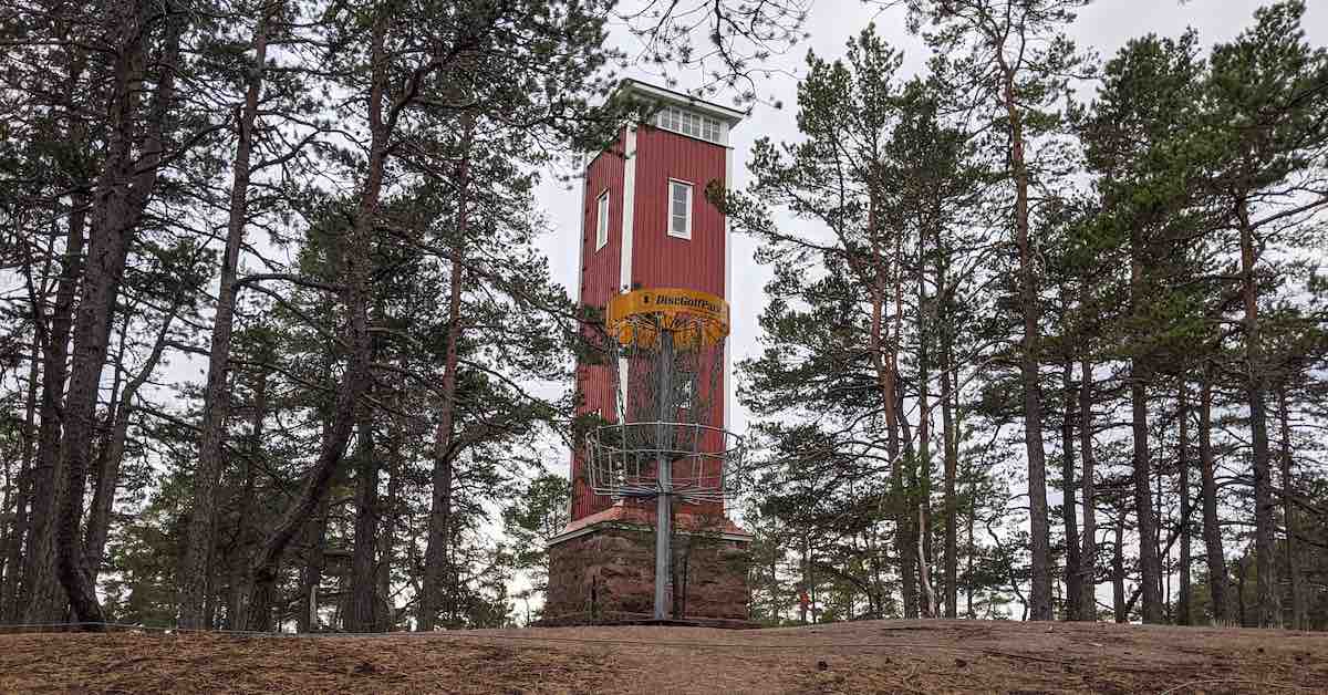 A yellow-banded disc golf basket in front of a very Scandinavian wooden tower painted white and red