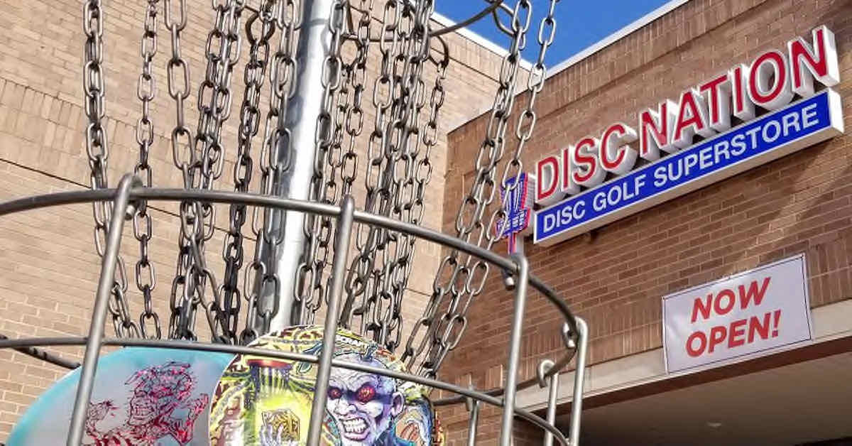 A sign saying "Disc Nation" on a brick building as seen through the chains of a disc golf basket