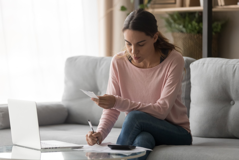 Woman sat on her sofa with the laptop open, she's using a calculator to work out some finances and making notes on paper.