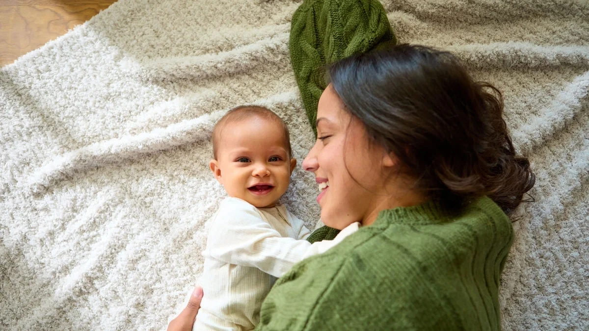 Parent lying on a soft blanket, smiling and cuddling a happy baby who is looking toward the camera.