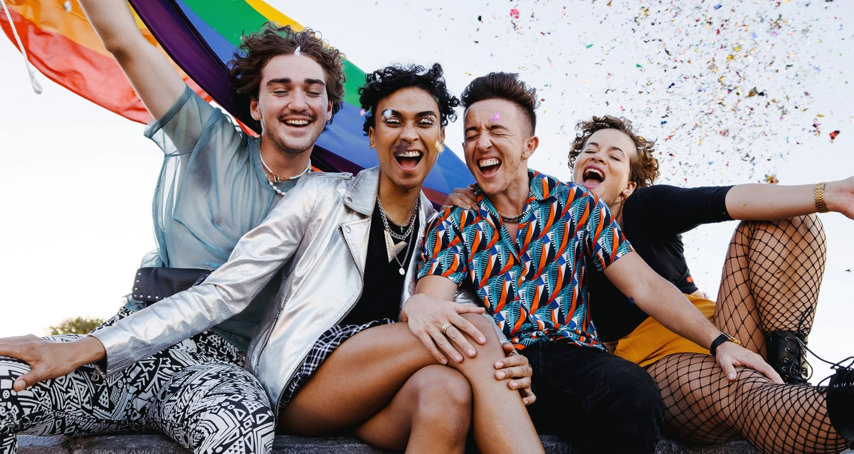 A group of queer folks laughing with a pride flag celebrating LGBTQ joy.