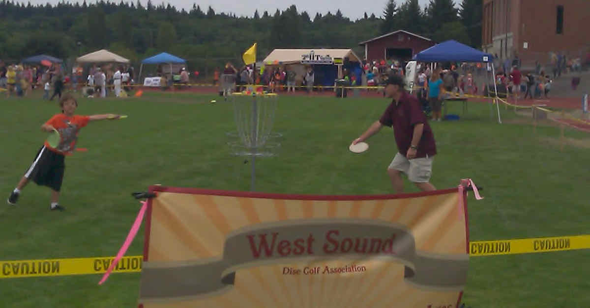 An older man and young child putt at a disc golf basket in a festival-like atmosphere