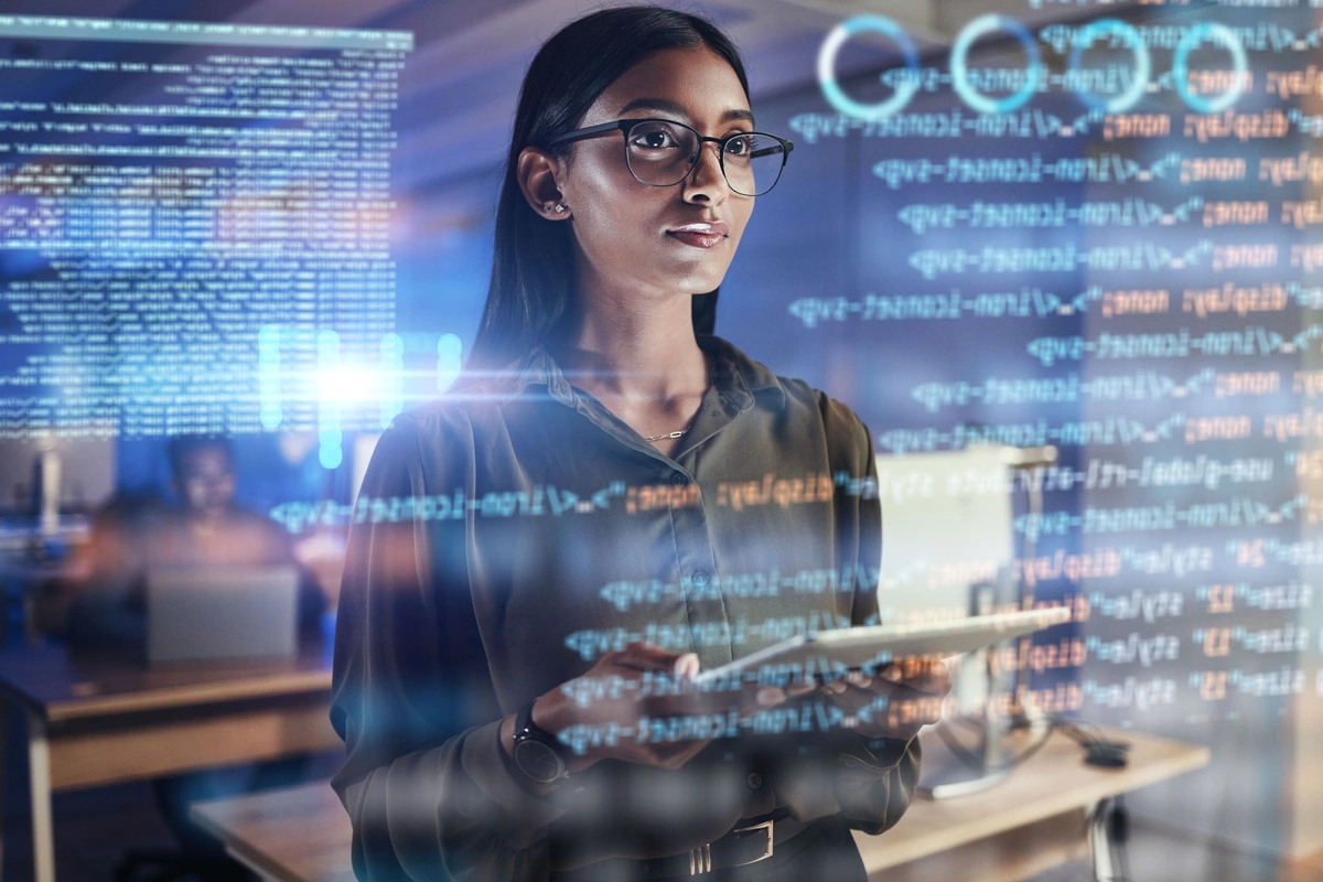 A woman stands in front of a transparent screen filled with lines of code, holding a tablet and interacting with the data. The background suggests an office environment with blurred details.