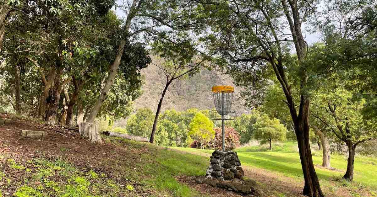Disc golf basket mounted on a small pile of stones. Hill in the background and surrounded by trees