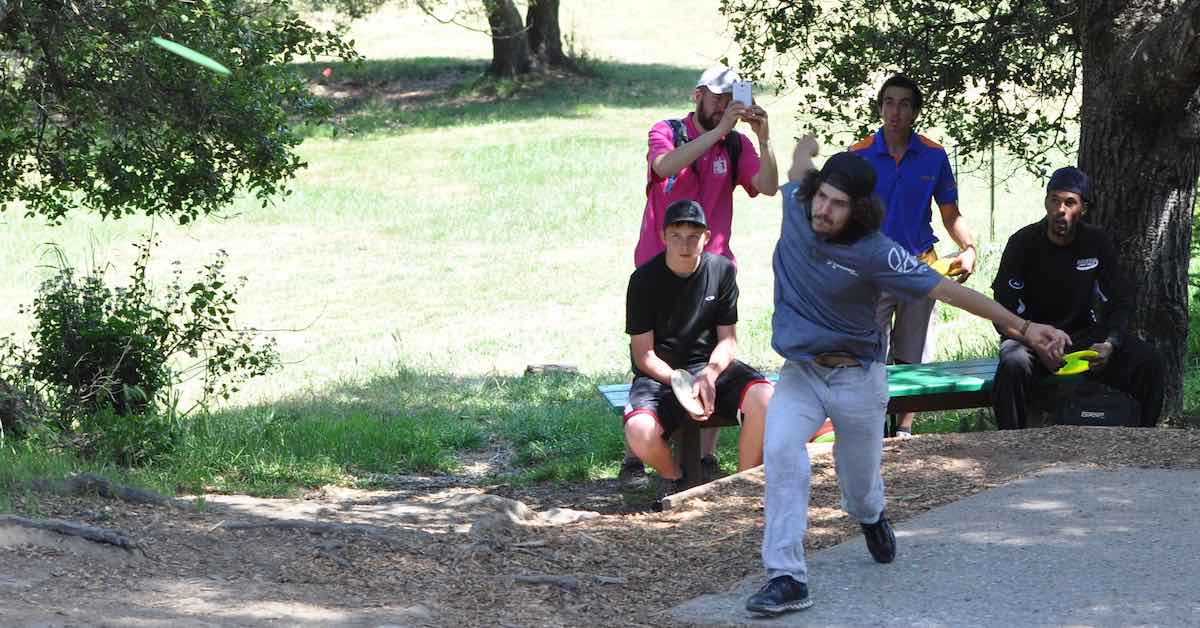 A young man on a disc golf tee pad just after releasing his disc. A group of four others look on