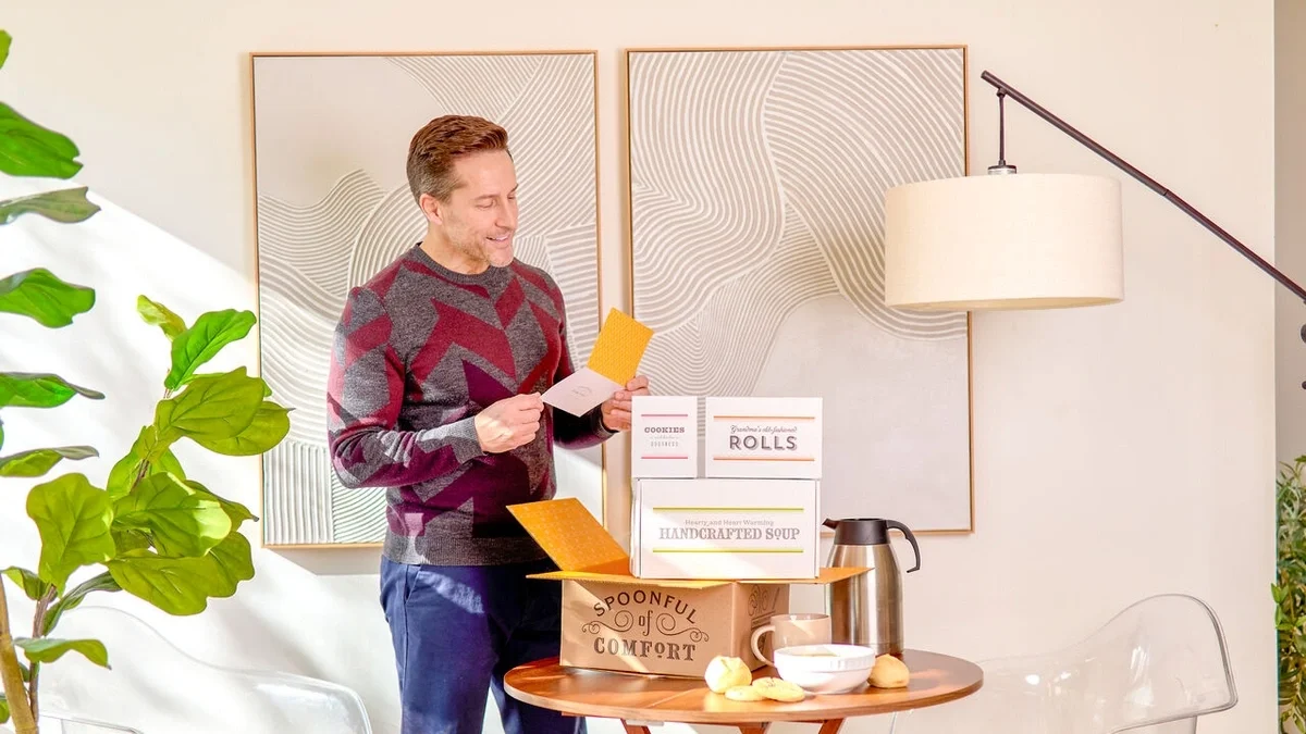 Man standing beside a table, smiling as he reads a greeting card after opening a Spoonful of Comfort gift box, with packaged soup, rolls, cookies, and a bowl of soup nearby.