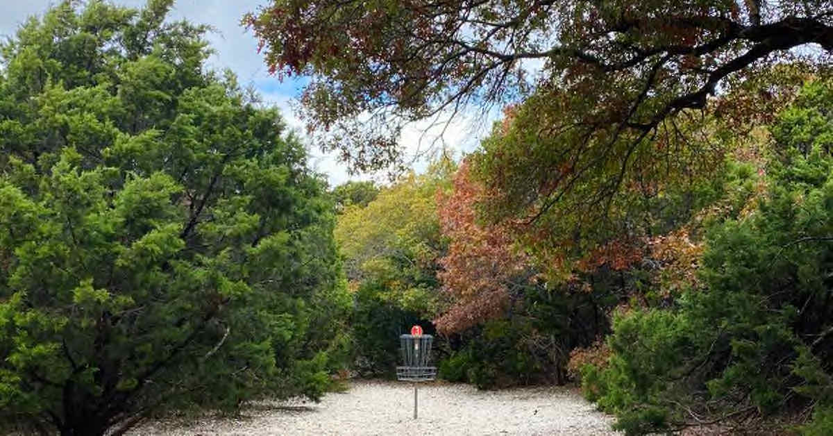 A disc golf basket on gravelly ground tightly surrounded by trees