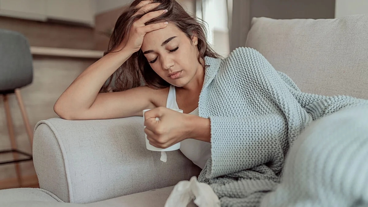 Person resting on a couch wrapped in a blanket, holding a mug of tea and touching their forehead while feeling under the weather.