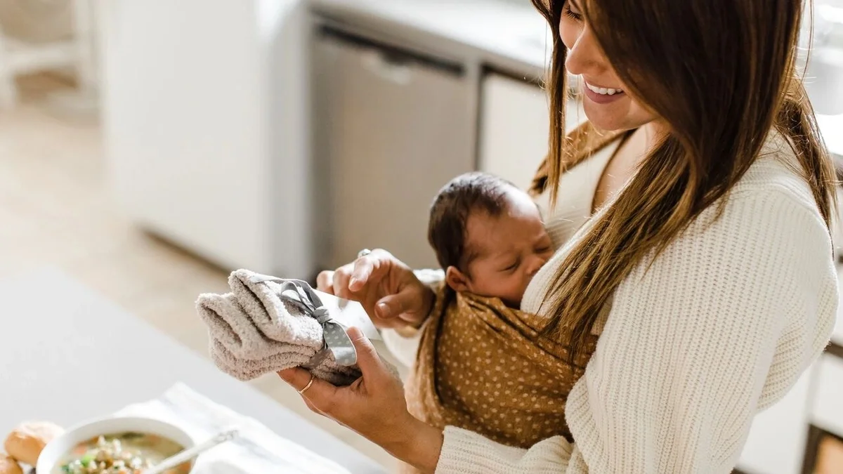 Mother smiling while holding a sleeping baby in a soft wrap and a pair of cozy socks, with a bowl of soup nearby.