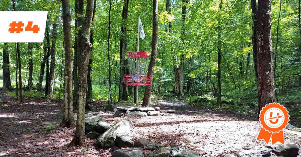A red disc golf basket in the woods with large rocks around it. #4 and UDisc logo in ribbon superimposed