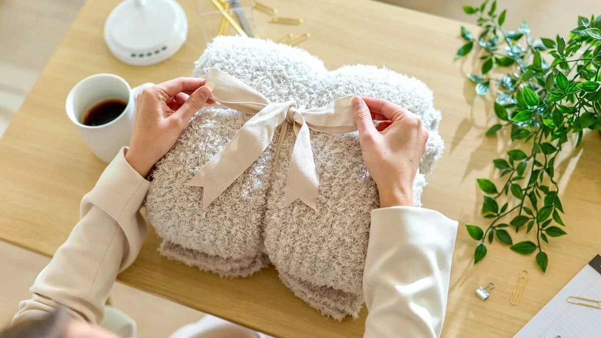 Overhead view of hands tying a satin ribbon around a soft, plush throw blanket on a wooden desk, with a mug and green plant nearby.
