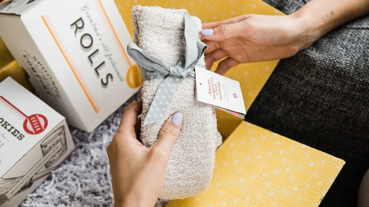 Close-up of hands holding a pair of cozy socks tied with a gray ribbon and gift tag, with Spoonful of Comfort boxes and yellow gift wrap in the background.