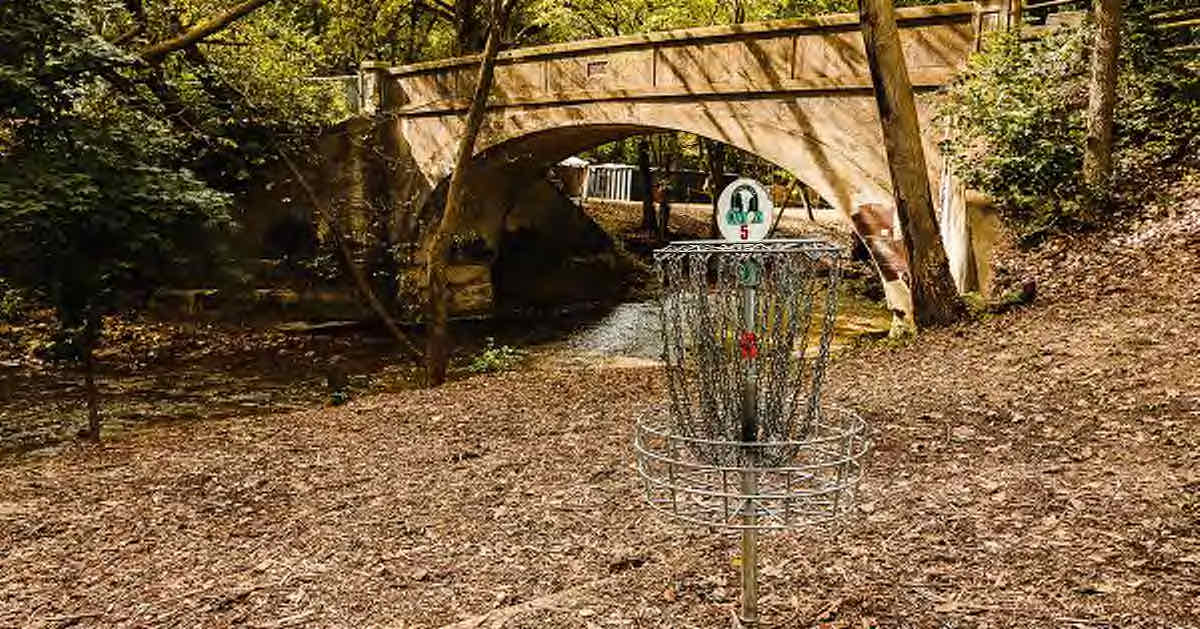 A disc golf basket in front of an old concrete bridge over a creek